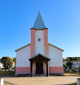 A church with a tall steeple and a cross at the top. The building is painted white with pink accents and has a front porch supported by dark wooden beams. The background is a clear blue sky, and there are houses and trees surrounding the church.