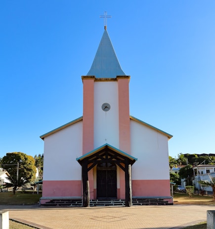 A church with a tall steeple and a cross at the top. The building is painted white with pink accents and has a front porch supported by dark wooden beams. The background is a clear blue sky, and there are houses and trees surrounding the church.