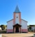 A church with a tall steeple and a cross at the top. The building is painted white with pink accents and has a front porch supported by dark wooden beams. The background is a clear blue sky, and there are houses and trees surrounding the church.