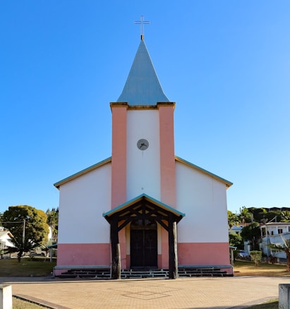A church with a tall steeple and a cross at the top. The building is painted white with pink accents and has a front porch supported by dark wooden beams. The background is a clear blue sky, and there are houses and trees surrounding the church.