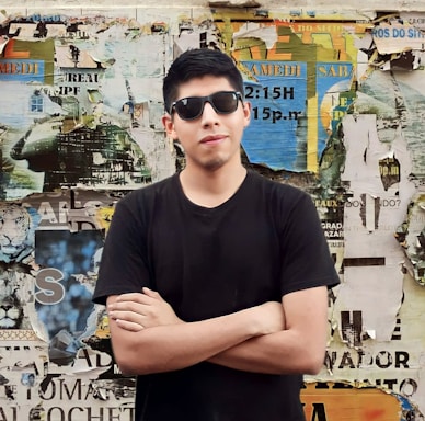 A person wearing sunglasses and a black t-shirt stands confidently with arms crossed in front of a wall covered with torn and layered colorful posters and advertisements.