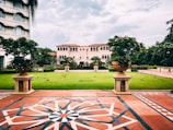 A landscaped garden area with a lush green lawn surrounded by a symmetrical arrangement of plants in large decorative pots. The scene is framed by a patterned tiled walkway in the foreground, with an ornate building featuring arches and multiple windows in the background. Birds are scattered on the grass, and a few are in flight.