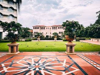 A landscaped garden area with a lush green lawn surrounded by a symmetrical arrangement of plants in large decorative pots. The scene is framed by a patterned tiled walkway in the foreground, with an ornate building featuring arches and multiple windows in the background. Birds are scattered on the grass, and a few are in flight.