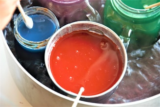 Various colored jars and containers filled with liquid dye are placed inside a larger silver pot. Each jar contains a wooden stick used for stirring. The colors of the dyes include vibrant red, blue, green, and purple.