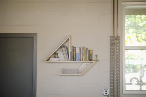 A minimalist triangular wall shelf holds a few books, magazines, and a small trophy. The shelves are mounted on a light-colored brick wall. To the left, there is a dark gray door, and to the right is a window with a view of greenery outside. Metal conduit pipes run vertically alongside the window.