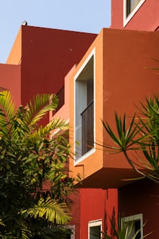A modern architectural exterior is highlighted by bold, angular shapes and vibrant red and orange tones. Large windows are accented by white frames, creating a stark contrast against the vibrant colors. Lush green foliage partially obscures parts of the structure, adding a natural element to the urban setting.
