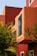 A modern architectural exterior is highlighted by bold, angular shapes and vibrant red and orange tones. Large windows are accented by white frames, creating a stark contrast against the vibrant colors. Lush green foliage partially obscures parts of the structure, adding a natural element to the urban setting.