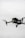 A black quadcopter drone with green lights is captured in mid-flight against a cloudy, gray sky. The drone has a camera mounted on the front and rotating propellers visible.