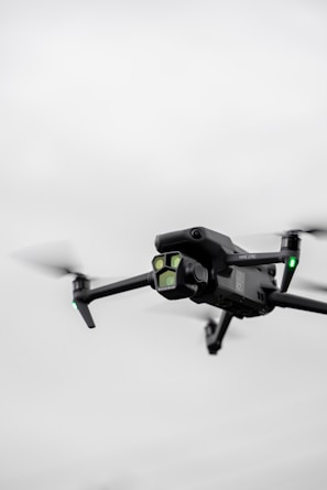 A black quadcopter drone with green lights is captured in mid-flight against a cloudy, gray sky. The drone has a camera mounted on the front and rotating propellers visible.