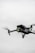 A black quadcopter drone with green lights is captured in mid-flight against a cloudy, gray sky. The drone has a camera mounted on the front and rotating propellers visible.