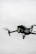 A black quadcopter drone with green lights is captured in mid-flight against a cloudy, gray sky. The drone has a camera mounted on the front and rotating propellers visible.
