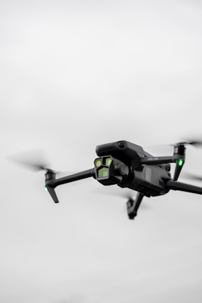 A black quadcopter drone with green lights is captured in mid-flight against a cloudy, gray sky. The drone has a camera mounted on the front and rotating propellers visible.
