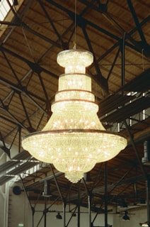 A large, ornate chandelier hangs from a high wooden ceiling, featuring multiple layers adorned with sparkling crystals and numerous light bulbs, casting a warm glow. The surrounding area includes metal beams and industrial fixtures.