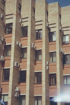 The image features a brick building facade with multiple air conditioning units installed on the walls between the windows. The building has a modern architectural design with uniform, rectangular windows. The brickwork alternates between lighter and darker shades, giving a textured appearance to the structure. There is a slight glare or light reflection visible at the bottom right corner.