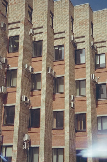The image features a brick building facade with multiple air conditioning units installed on the walls between the windows. The building has a modern architectural design with uniform, rectangular windows. The brickwork alternates between lighter and darker shades, giving a textured appearance to the structure. There is a slight glare or light reflection visible at the bottom right corner.