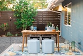 A cozy outdoor workspace set in a backyard features a wooden table with a laptop, books, glassware, and decorative items. Two poufs serve as seating on a patterned outdoor rug. String lights hang above the area, creating a warm ambiance. In the background, a wooden fence and greenery, including a lemon tree, add a natural touch.