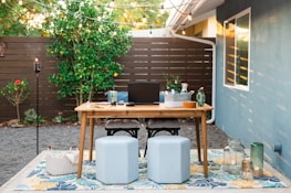 A cozy outdoor workspace set in a backyard features a wooden table with a laptop, books, glassware, and decorative items. Two poufs serve as seating on a patterned outdoor rug. String lights hang above the area, creating a warm ambiance. In the background, a wooden fence and greenery, including a lemon tree, add a natural touch.
