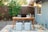 A cozy outdoor workspace set in a backyard features a wooden table with a laptop, books, glassware, and decorative items. Two poufs serve as seating on a patterned outdoor rug. String lights hang above the area, creating a warm ambiance. In the background, a wooden fence and greenery, including a lemon tree, add a natural touch.