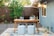 A cozy outdoor workspace set in a backyard features a wooden table with a laptop, books, glassware, and decorative items. Two poufs serve as seating on a patterned outdoor rug. String lights hang above the area, creating a warm ambiance. In the background, a wooden fence and greenery, including a lemon tree, add a natural touch.