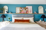 A well-decorated bedroom features a bed with a gray headboard, off-white bedding, and a prominent burnt orange accent pillow. On either side, there are wooden nightstands with black lamps and small décor items. A teal blue wall serves as a backdrop, adorned with colorful framed art, mirrors with rounded tops, and small plants.
