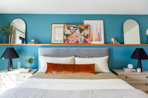 A well-decorated bedroom features a bed with a gray headboard, off-white bedding, and a prominent burnt orange accent pillow. On either side, there are wooden nightstands with black lamps and small décor items. A teal blue wall serves as a backdrop, adorned with colorful framed art, mirrors with rounded tops, and small plants.