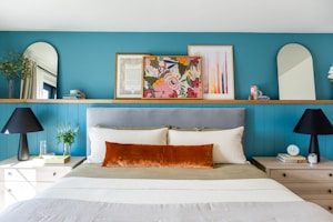 A well-decorated bedroom features a bed with a gray headboard, off-white bedding, and a prominent burnt orange accent pillow. On either side, there are wooden nightstands with black lamps and small d&eacute;cor items. A teal blue wall serves as a backdrop, adorned with colorful framed art, mirrors with rounded tops, and small plants.