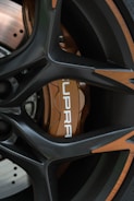 Close-up of a car wheel featuring a sporty black alloy rim with bronze accents. The brake caliper is visible, painted in a metallic bronze shade with the word 'SUPRA' printed in white. The disc brake is partially visible with a polished finish.