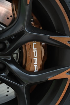 Close-up of a car wheel featuring a sporty black alloy rim with bronze accents. The brake caliper is visible, painted in a metallic bronze shade with the word 'SUPRA' printed in white. The disc brake is partially visible with a polished finish.