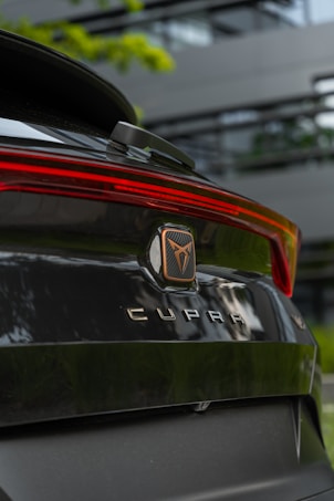 The rear view of a black car with the brand name 'CUPRA' prominently displayed, showcasing a sleek and modern design. The background includes blurred greenery and a modern building, which adds to the contemporary feel.
