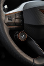 A close-up view of a car's steering wheel and part of the dashboard. The steering wheel has a textured, perforated surface, and several buttons, including ones for setting cruise control and adjusting volume. The dashboard features a carbon fiber-like texture and a logo in the center.