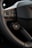 A close-up view of a car's steering wheel and part of the dashboard. The steering wheel has a textured, perforated surface, and several buttons, including ones for setting cruise control and adjusting volume. The dashboard features a carbon fiber-like texture and a logo in the center.