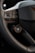 A close-up view of a car's steering wheel and part of the dashboard. The steering wheel has a textured, perforated surface, and several buttons, including ones for setting cruise control and adjusting volume. The dashboard features a carbon fiber-like texture and a logo in the center.