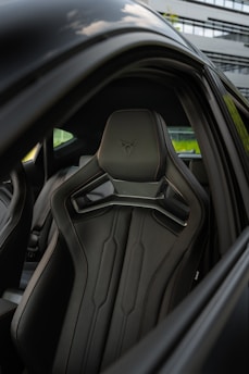 A close-up view of a sleek, modern car interior featuring a stylish black leather seat with distinct contours and stitching. The seat has a sporty design with an embedded logo on the headrest. The surroundings show part of the car door and window, with a blurred exterior building in the background.