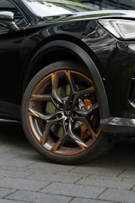 Close-up of a car wheel featuring a stylish alloy rim with orange and silver accents. The wheel is mounted on a dark-colored vehicle, parked on a tiled surface. The design of the rim is modern and sharp, showcasing intricate patterns.