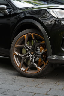 Close-up of a car wheel featuring a stylish alloy rim with orange and silver accents. The wheel is mounted on a dark-colored vehicle, parked on a tiled surface. The design of the rim is modern and sharp, showcasing intricate patterns.