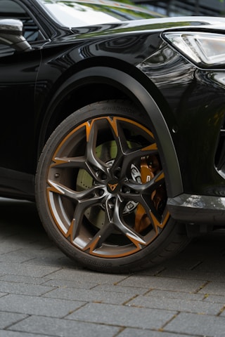 Close-up of a car wheel featuring a stylish alloy rim with orange and silver accents. The wheel is mounted on a dark-colored vehicle, parked on a tiled surface. The design of the rim is modern and sharp, showcasing intricate patterns.