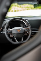 A detailed view of a car's steering wheel with a modern, sleek design. The steering wheel features multiple control buttons and a prominent logo at the center. The background shows part of the car's dashboard and a glimpse of the outside.