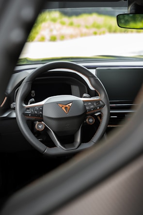 A detailed view of a car's steering wheel with a modern, sleek design. The steering wheel features multiple control buttons and a prominent logo at the center. The background shows part of the car's dashboard and a glimpse of the outside.