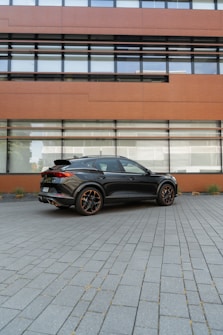 A sleek, modern black car is parked on a paved area in front of a contemporary building with large windows. The building has a reddish-brown facade, contrasting with the car's glossy finish.