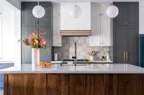 A modern kitchen with a large white marble island topped with a wooden base. There are three pendant lights hanging from the ceiling. A vase with colorful flowers and a bowl of oranges are placed on the island. The backsplash features a geometric pattern, and there is a stove with utensils and containers neatly arranged on the counter.