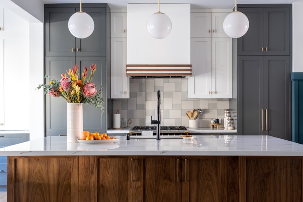 A modern kitchen with a large white marble island topped with a wooden base. There are three pendant lights hanging from the ceiling. A vase with colorful flowers and a bowl of oranges are placed on the island. The backsplash features a geometric pattern, and there is a stove with utensils and containers neatly arranged on the counter.