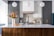 A modern kitchen with a large white marble island topped with a wooden base. There are three pendant lights hanging from the ceiling. A vase with colorful flowers and a bowl of oranges are placed on the island. The backsplash features a geometric pattern, and there is a stove with utensils and containers neatly arranged on the counter.