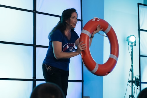 A woman holding an orange life preserver stands in front of a backdrop consisting of a grid of illuminated panels that give off a cool blue light. She is wearing a blue shirt and a microphone headset, suggesting she may be giving a presentation or conducting a demonstration.
