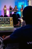 A person operates a sound mixing or audio control board, with multiple people on a stage in the background. The stage is illuminated with colorful lights, creating an atmospheric effect. The focus is on the technical setup, but there is a clear view of the performers in the background.