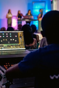 A person operates a sound mixing or audio control board, with multiple people on a stage in the background. The stage is illuminated with colorful lights, creating an atmospheric effect. The focus is on the technical setup, but there is a clear view of the performers in the background.