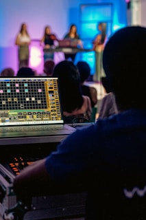 A person operates a sound mixing or audio control board, with multiple people on a stage in the background. The stage is illuminated with colorful lights, creating an atmospheric effect. The focus is on the technical setup, but there is a clear view of the performers in the background.
