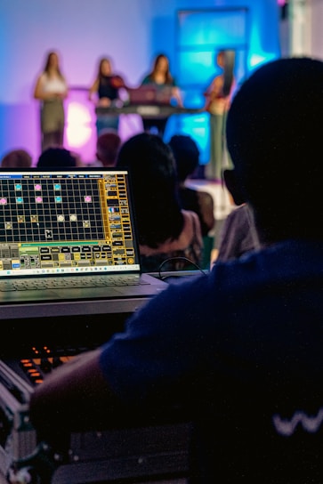 A person operates a sound mixing or audio control board, with multiple people on a stage in the background. The stage is illuminated with colorful lights, creating an atmospheric effect. The focus is on the technical setup, but there is a clear view of the performers in the background.
