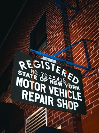 A sign reads 'Registered Motor Vehicle Repair Shop, State of New York' and is mounted against a red brick wall. The scene is illuminated by ambient lighting, casting shadows on the text and the wall.