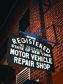 A sign reads 'Registered Motor Vehicle Repair Shop, State of New York' and is mounted against a red brick wall. The scene is illuminated by ambient lighting, casting shadows on the text and the wall.