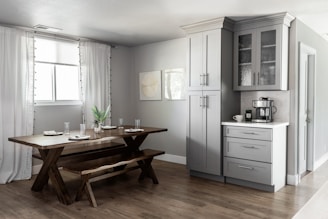 A modern kitchen and dining area featuring light gray cabinets with glass-paneled doors, a dark wooden dining table with matching benches, and decoratively arranged dishes and glasses. A coffee machine sits on a white countertop, and a potted plant is placed in the center of the dining table. Soft, natural light filters through white draped curtains covering a window.